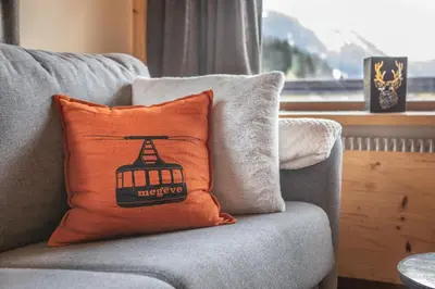 an orange pillow on a gray couch with a window at Les Corneilles - Appartement in Megève