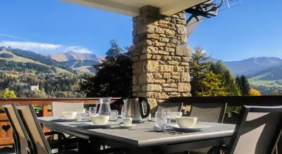 a table with plates and glasses on a balcony with mountains at Chalet Orchidee Megeve sur les pistes in Megève