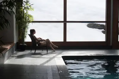 a woman sitting in a chair by a window next to a swimming pool at CGH Résidences & Spas Les Chalets de Jouvence in Les Carroz d'Araches
